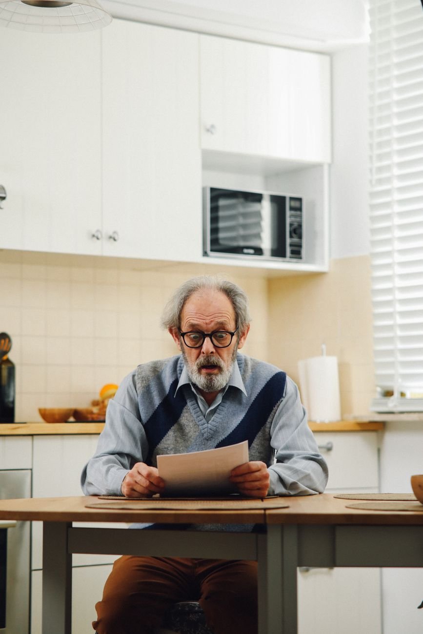 Senior man with eyeglasses reading a document at kitchen table, reflecting surprise.